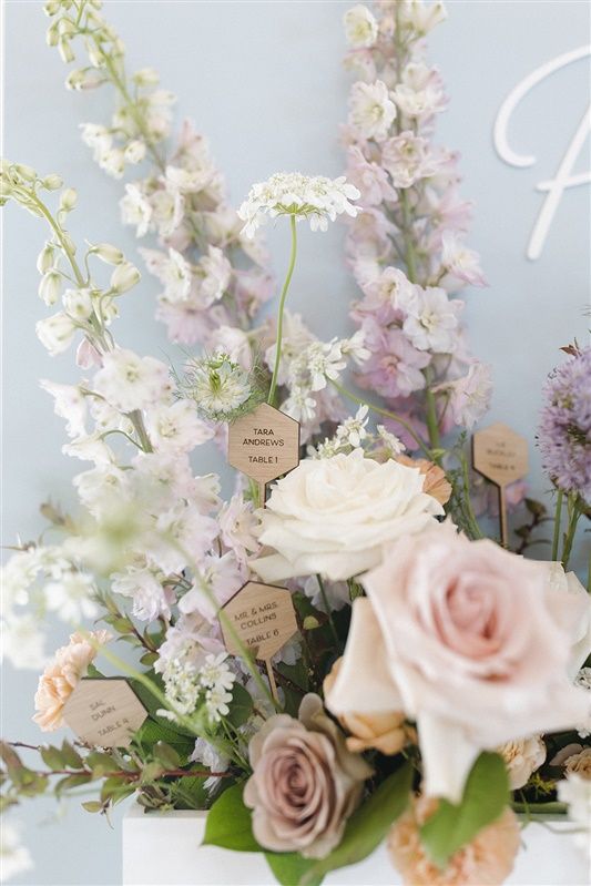 Elegant pastel floral arrangement with pink roses, delicate larkspur, and small decorative name cards on light gray background.