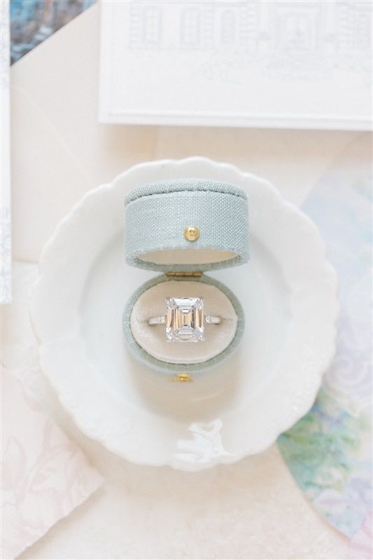 A stunning emerald-cut diamond engagement ring nestled in a pale blue velvet ring box on a white scalloped dish.