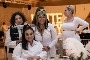 Wedding guests dressed in white attire gather around a decorated table at an indoor reception venue.