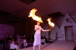 Fire dancer performs with dual flame props on a white stage with dramatic purple lighting.