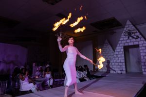 Fire dancer in white dress performs on stage with multiple flaming props in purple-lit venue.