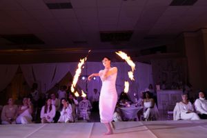 Fire performer spins flaming torches while dancing barefoot on a white stage.