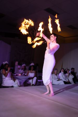 Performer in white dress juggles multiple fire props during an indoor stage show.