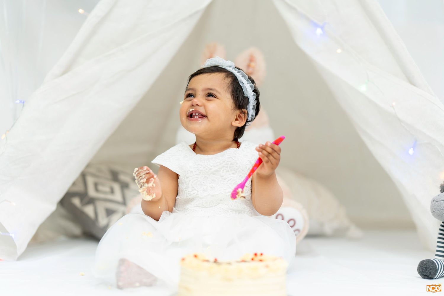 Young child in white dress sitting in teepee tent with birthday cake.