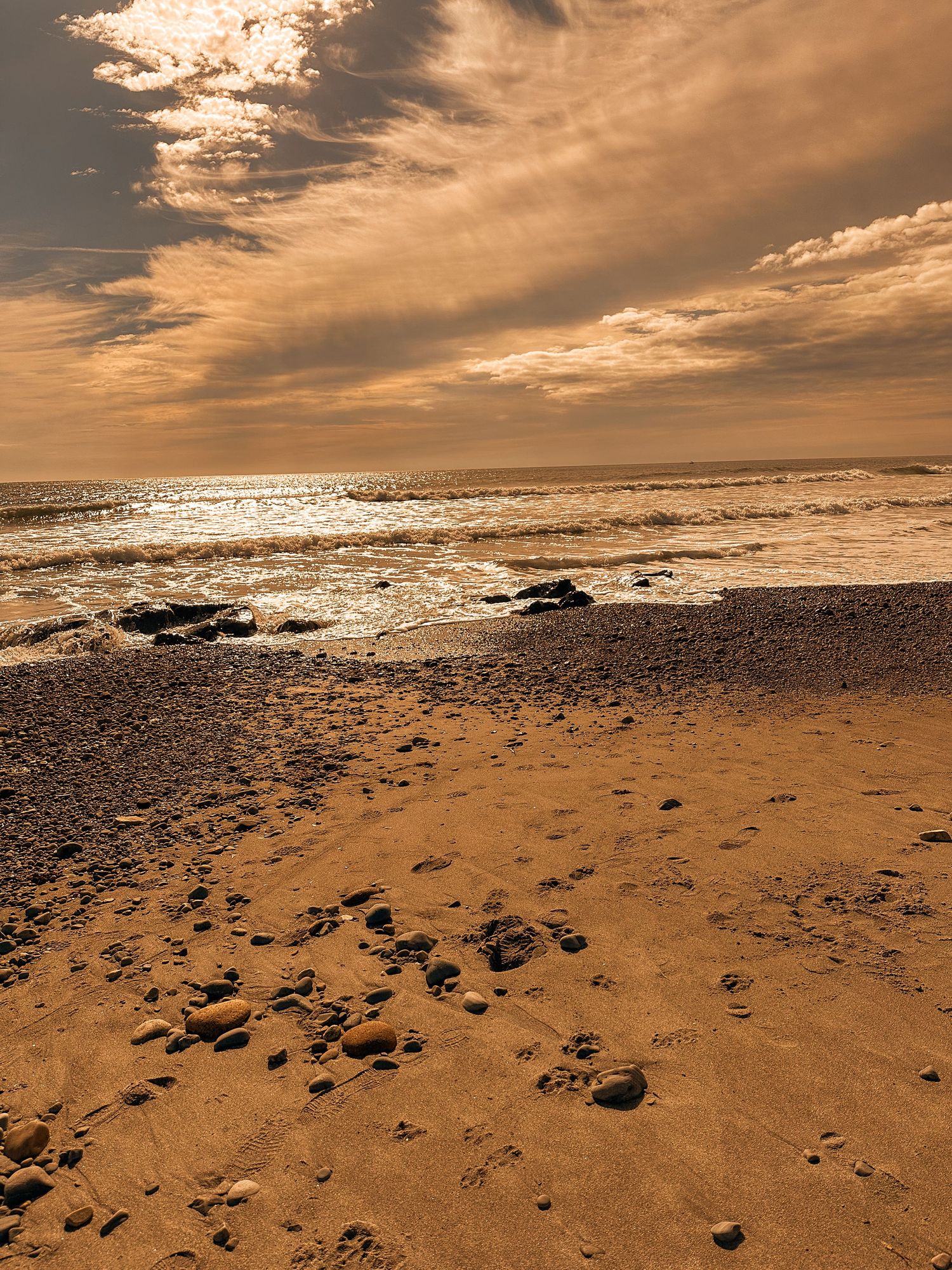 Dramatic seascape at sunset with rocky beach and golden clouds reflecting on water.