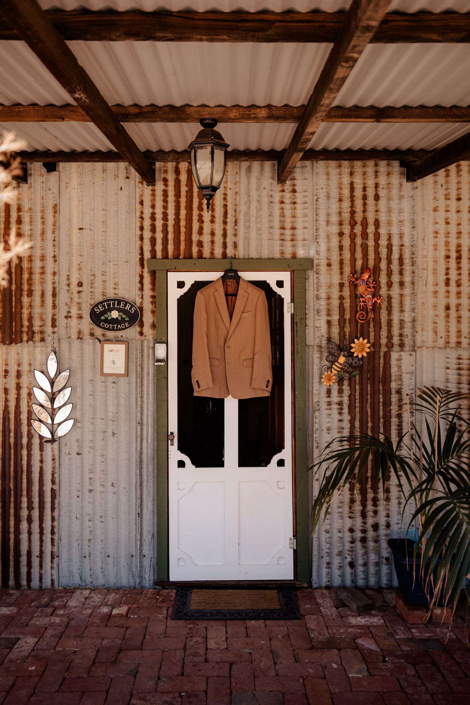A tan suit jacket hangs on a white screen door against a rustic corrugated metal wall with decorative elements.