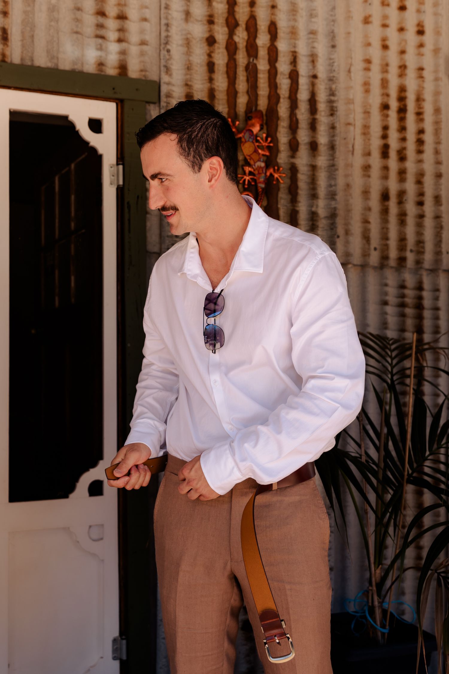 A groomsman in a crisp white shirt and tan pants smiles while standing on a rustic porch.