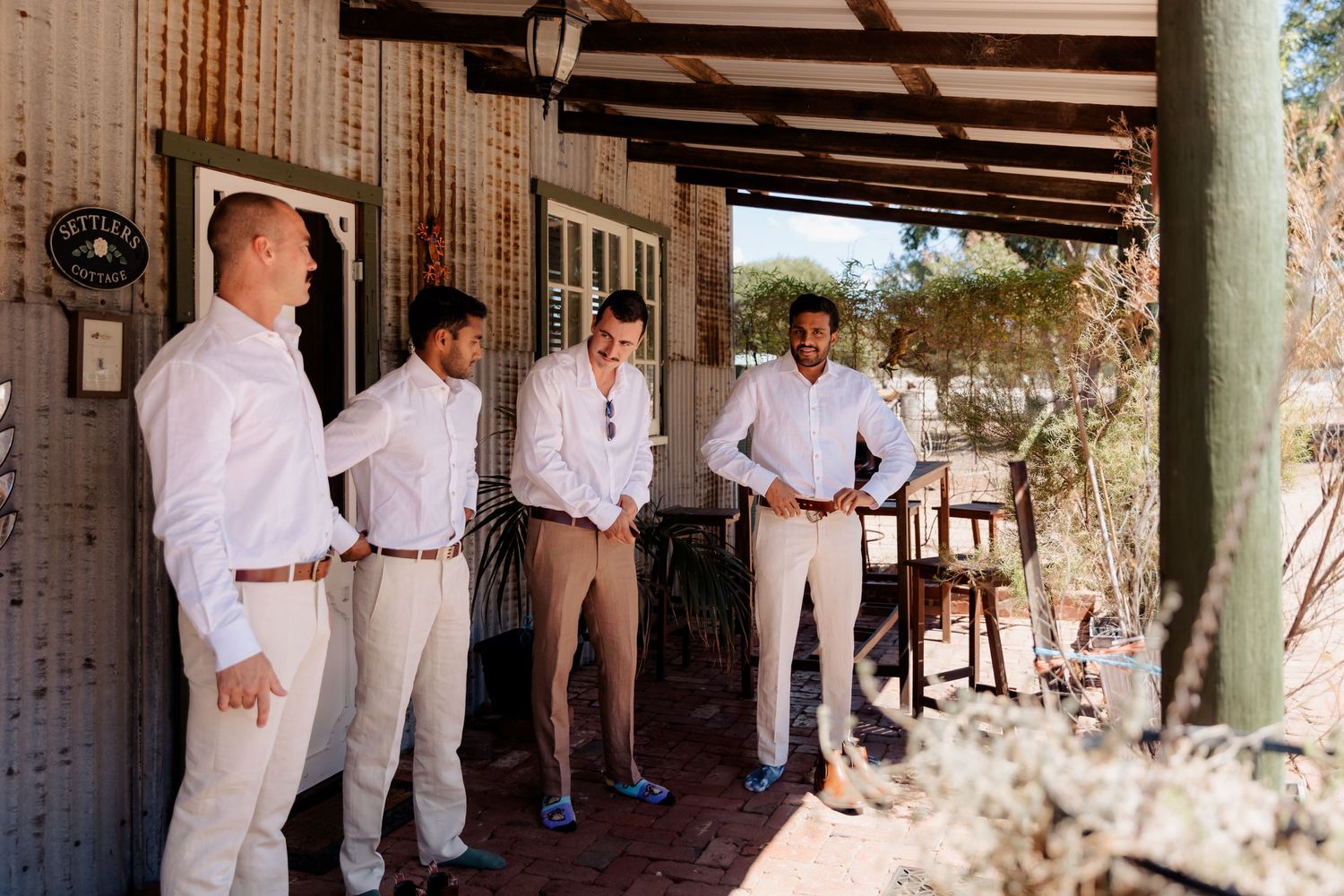 Wedding party members lined up on a covered porch wearing coordinated white shirts and light colored pants.