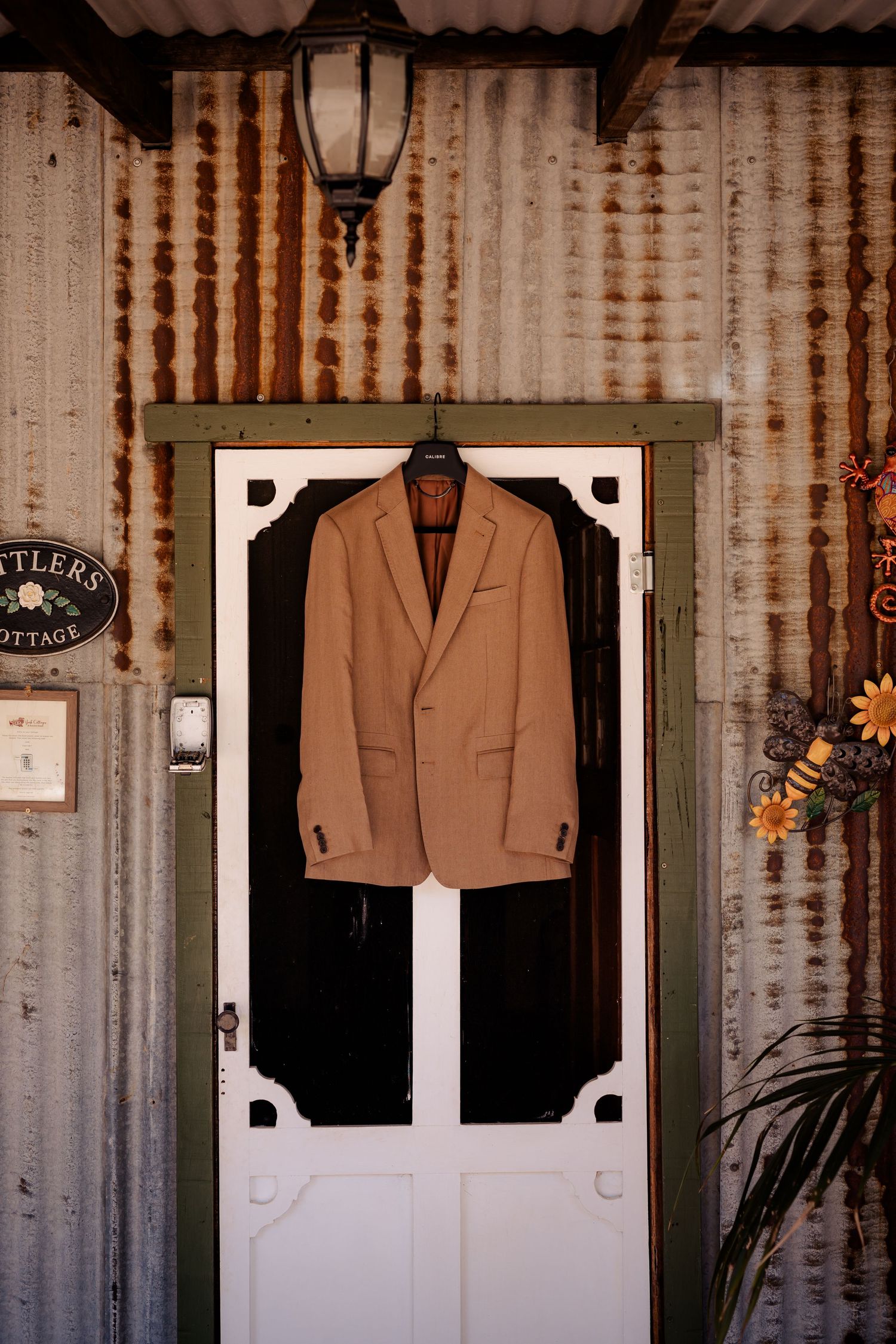 Tan suit jacket displayed on white screen door with vintage corrugated metal wall background and lantern above.