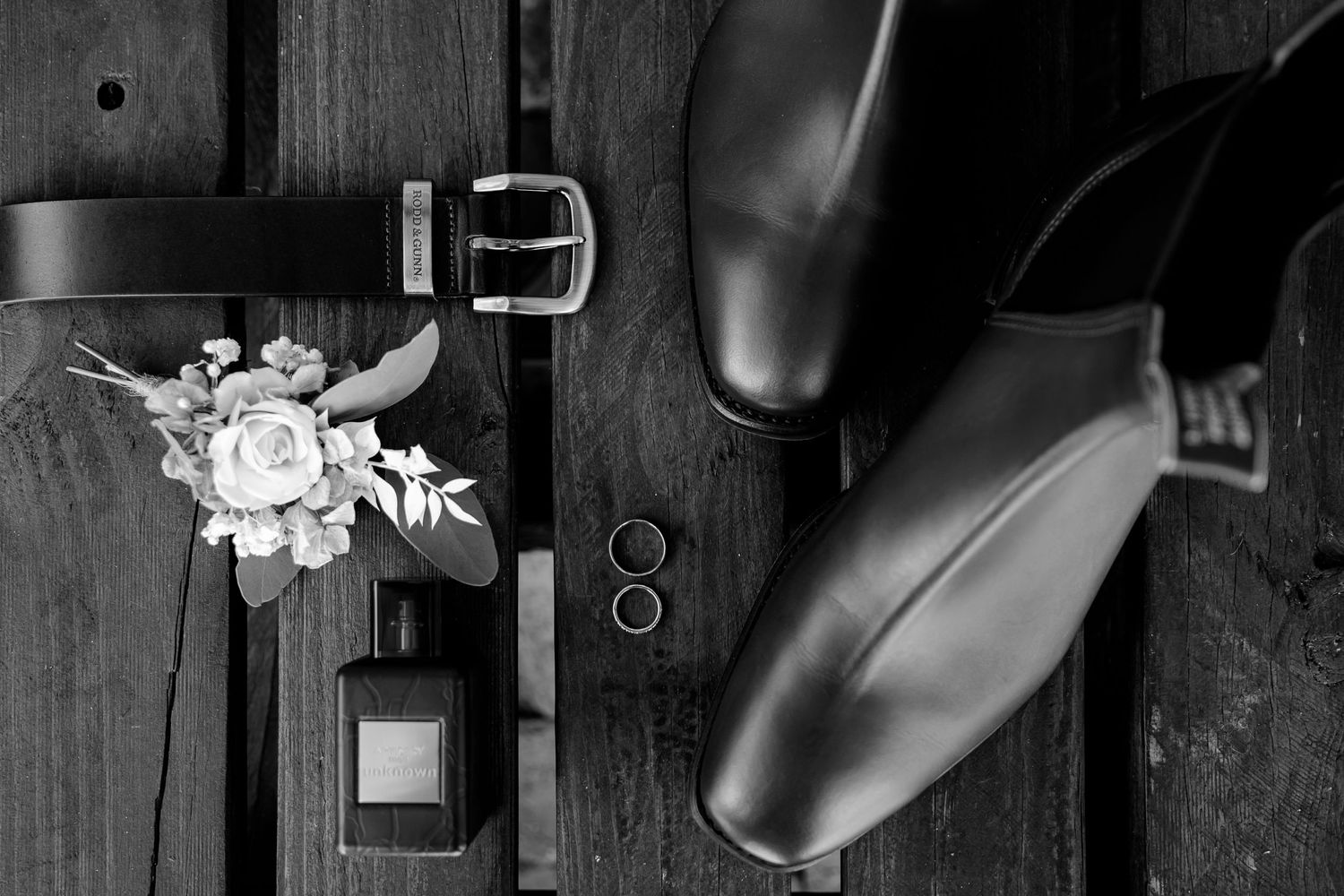 Wedding details including dress shoes, floral accent and rings arranged on dark wooden surface.