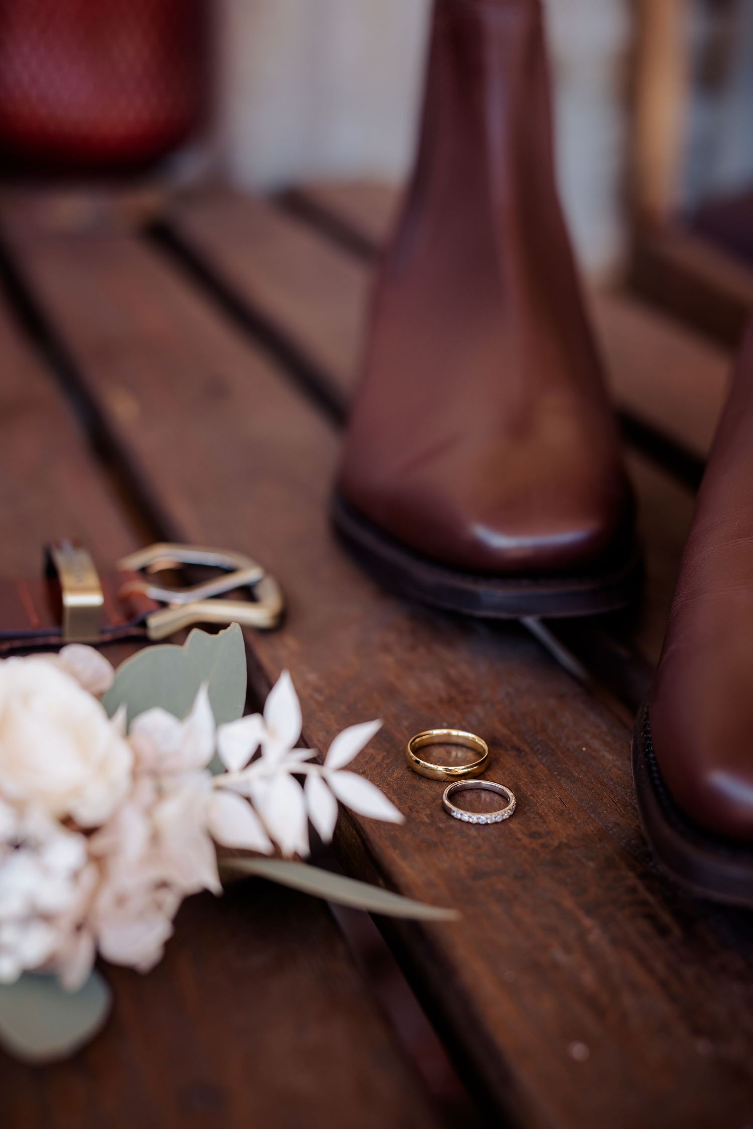 Brown leather boots with wedding rings and delicate floral arrangement on wooden surface.