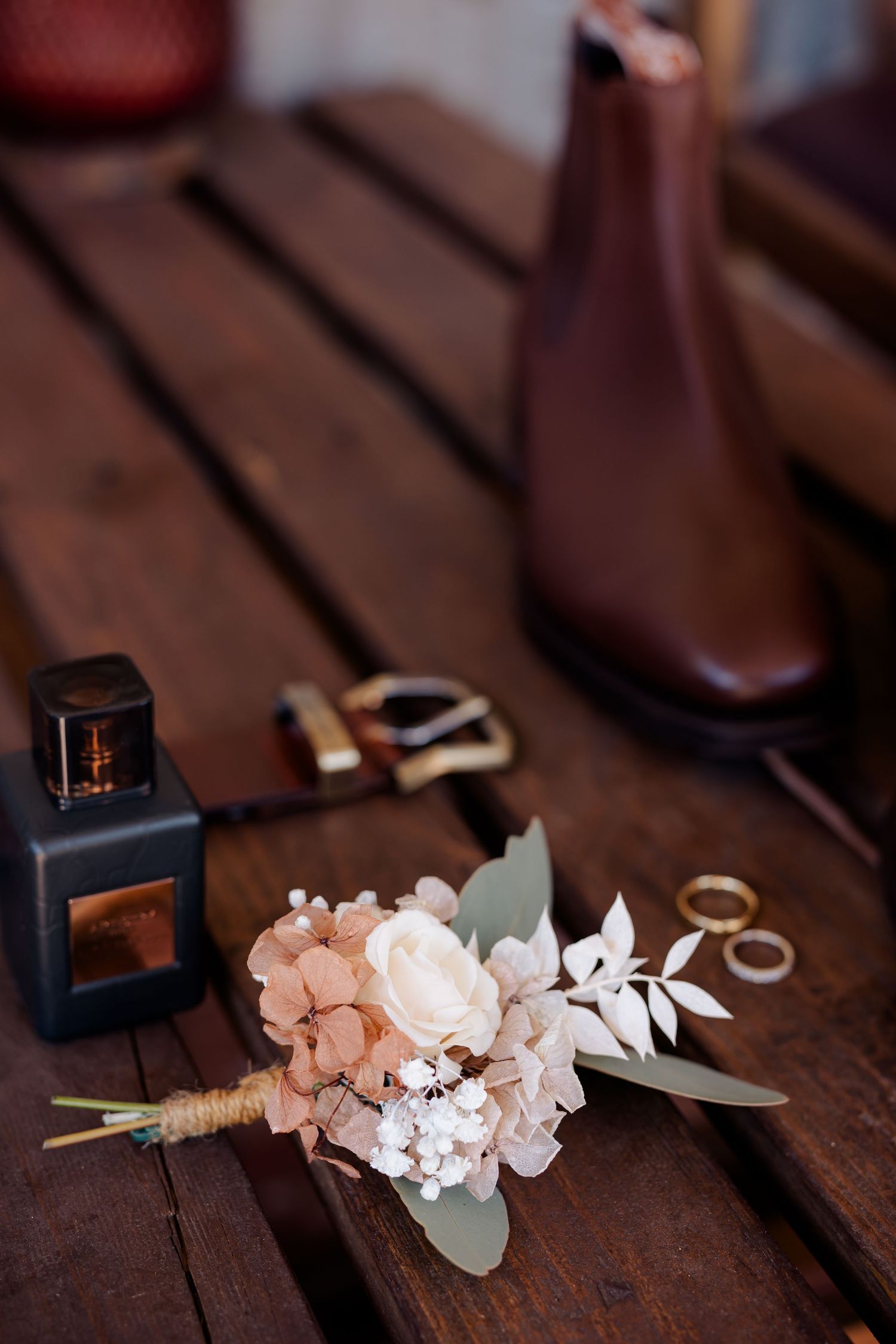 Wedding accessories including perfume, flowers, rings and brown boots arranged on wooden surface.