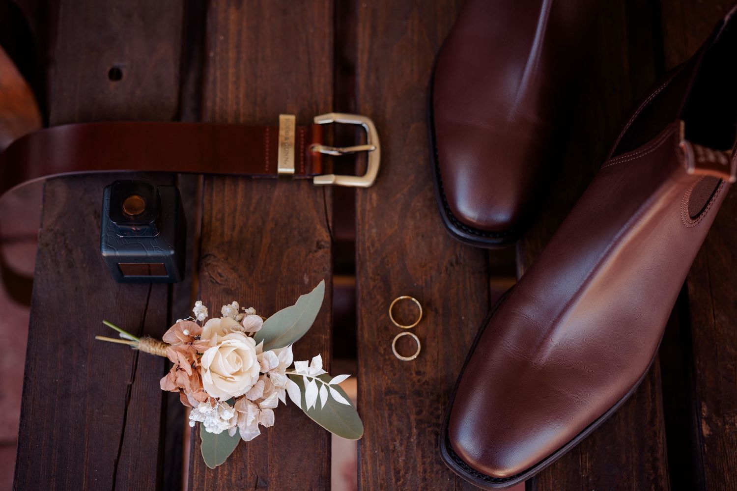 Wedding details featuring brown leather shoes, floral boutonniere, and rings on dark wood.