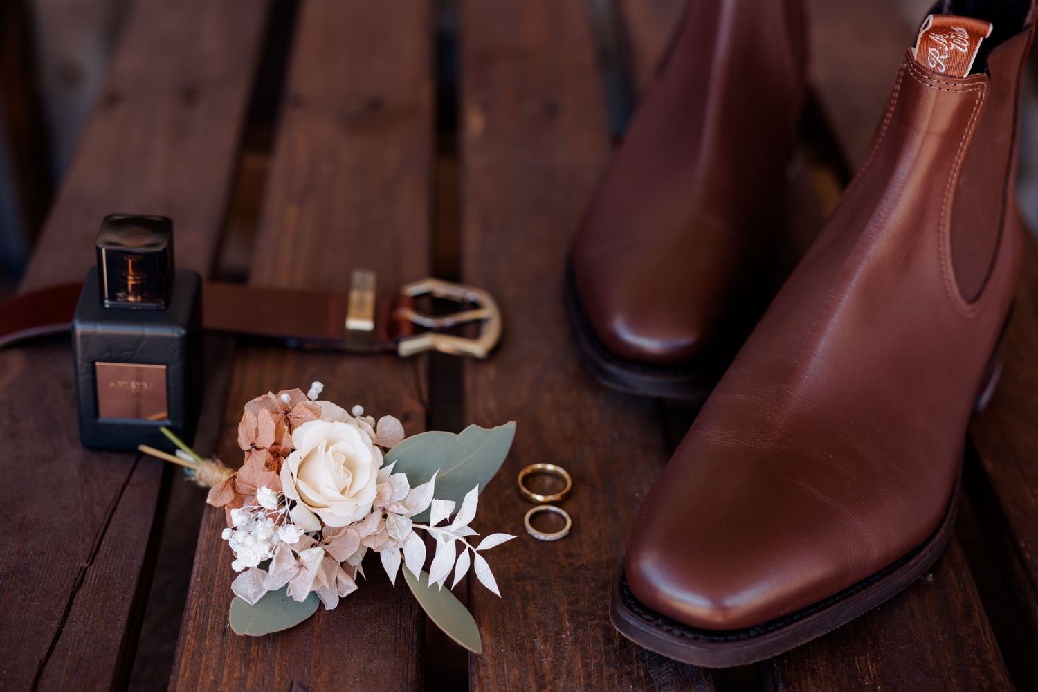 Wedding accessories including leather boots, cologne, floral arrangement and rings styled on wooden surface.