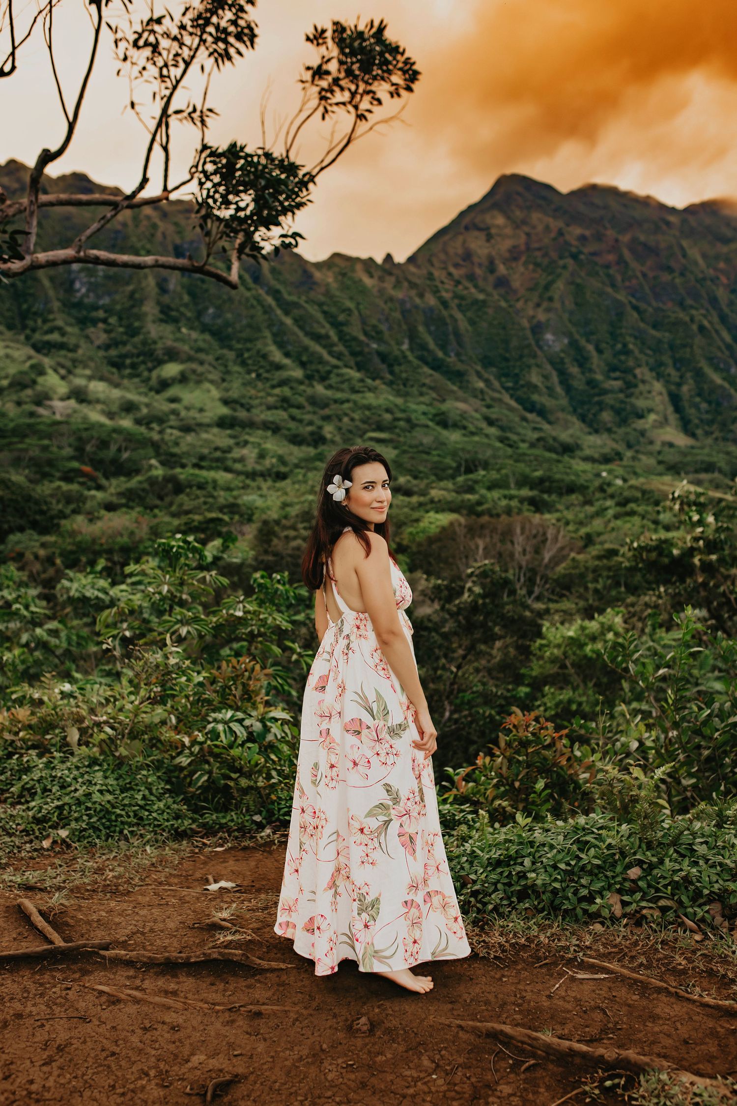 Woman in floral dress poses against mountainous Hawaiian landscape at sunset.