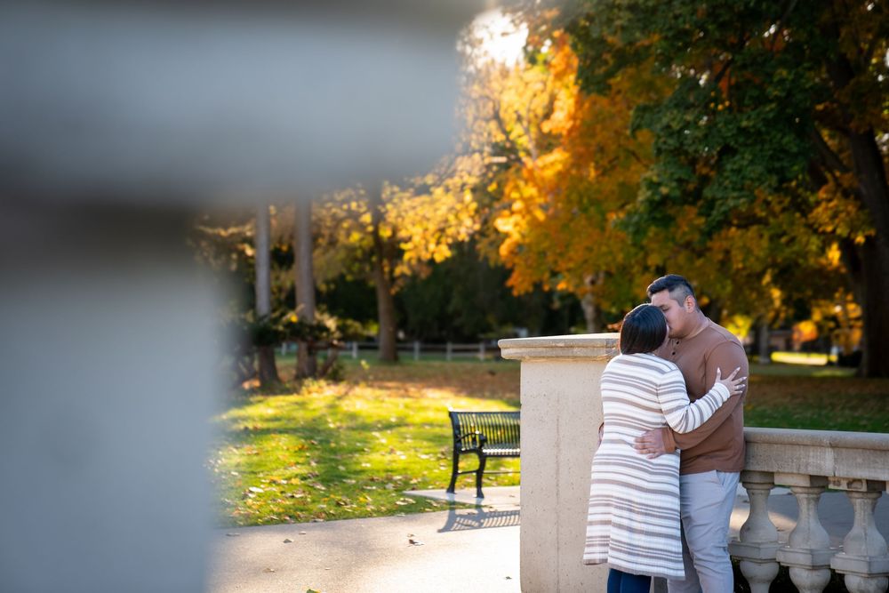A couple embraces and kisses while standing next to a white pillar in a sunlit outdoor park setting.