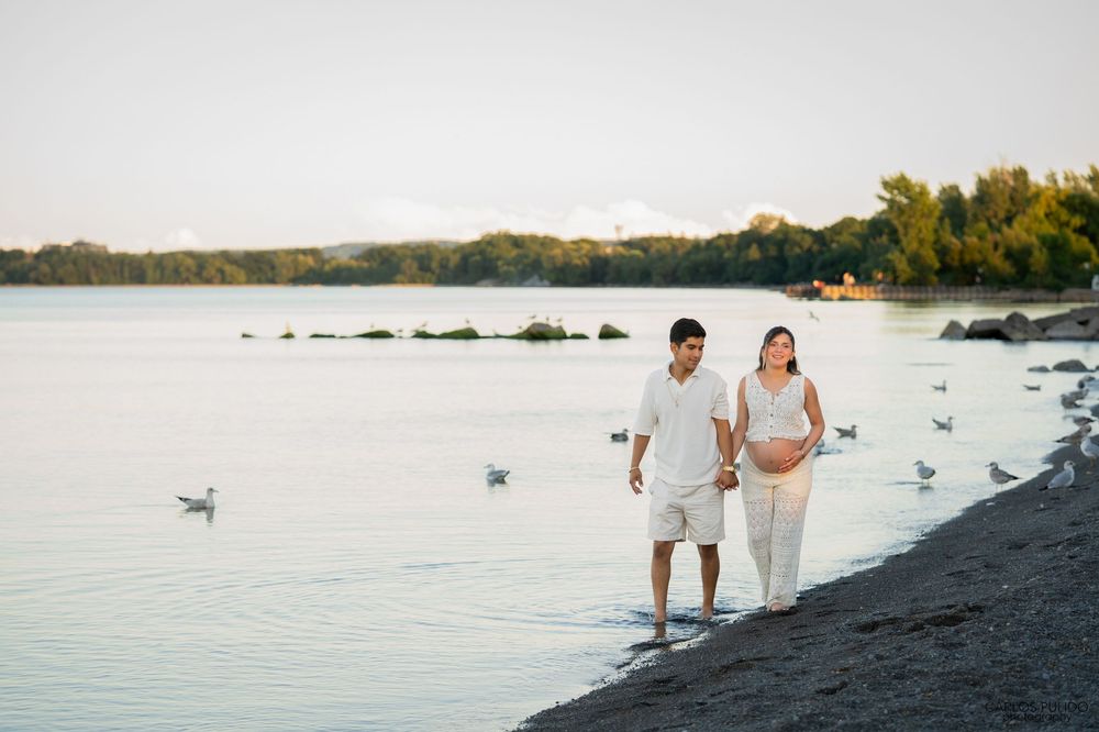A couple walks along a tranquil beach at sunset with seagulls floating on the calm water nearby.