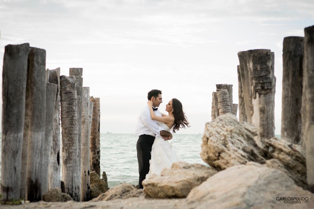 A romantic couple embraces on a rocky beach with weathered wooden pilings and ocean waves in the background.