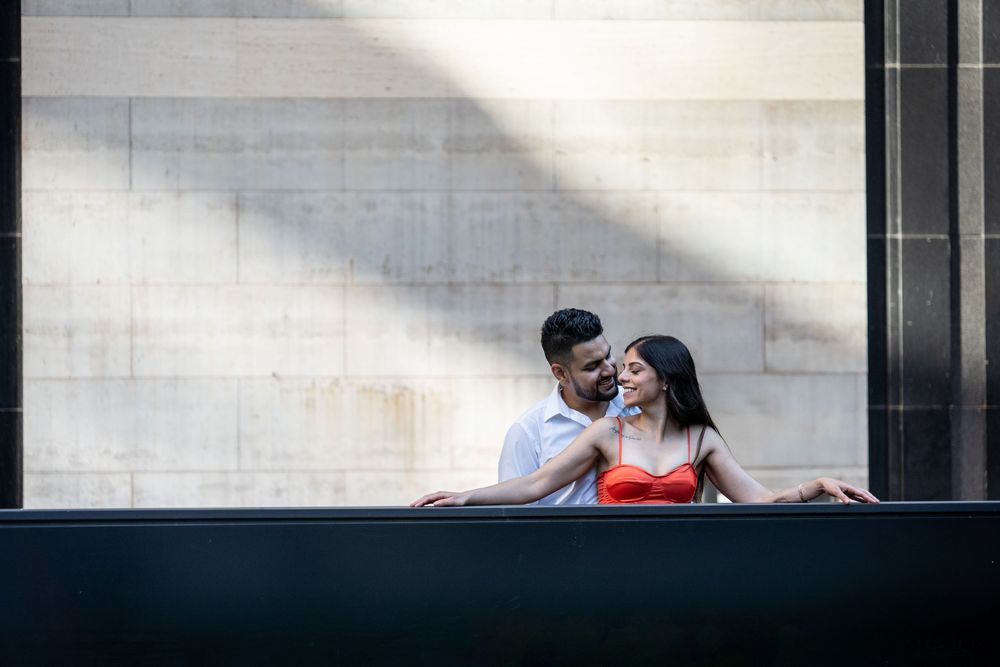 A couple shares a romantic moment while sitting on a bench in front of a modern building with glass windows.