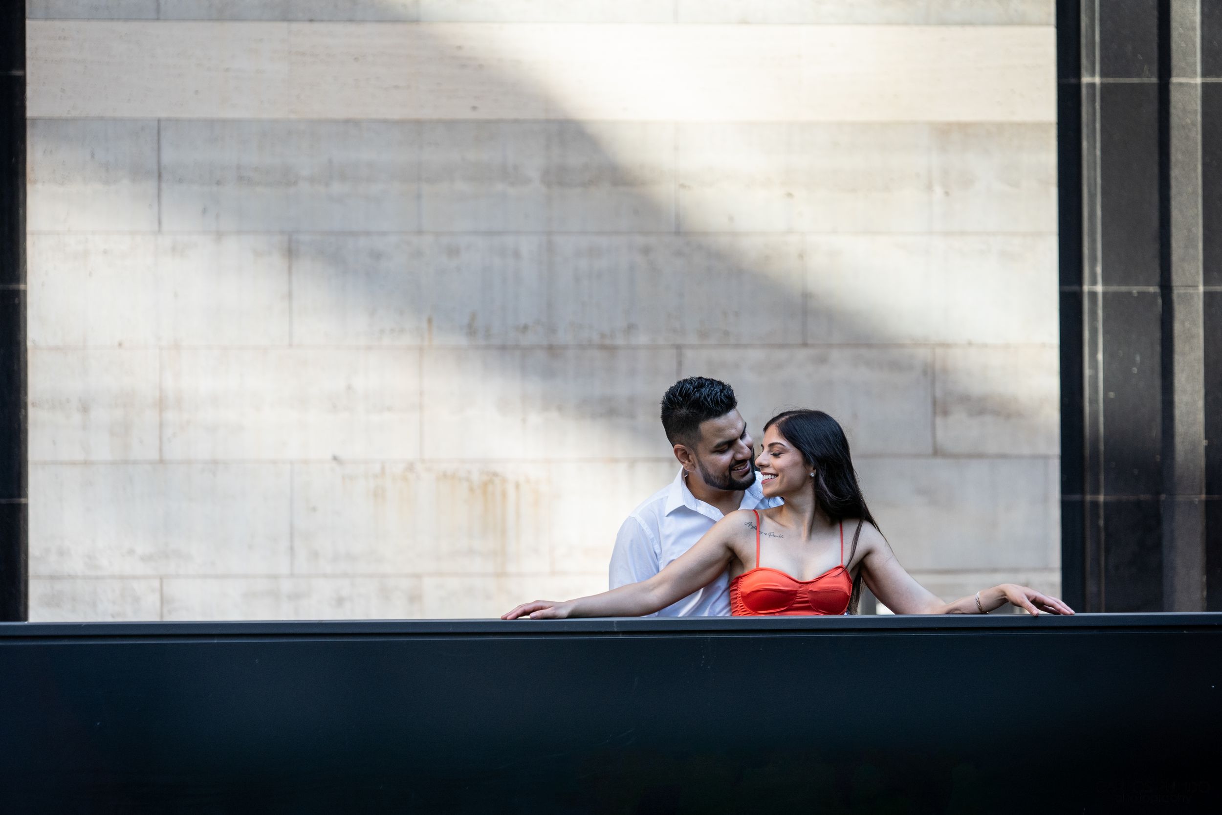 A couple shares a romantic moment while sitting on a bench in front of a modern building with glass windows.