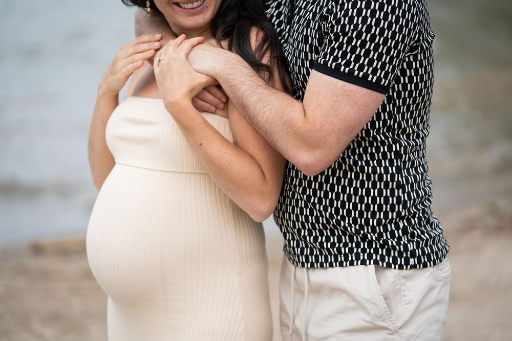 A close-up maternity photo shows a pregnant woman in a cream dress being embraced from behind on a beach.