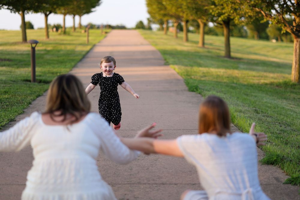 A child runs joyfully down a tree-lined path towards two people with outstretched arms at sunset.