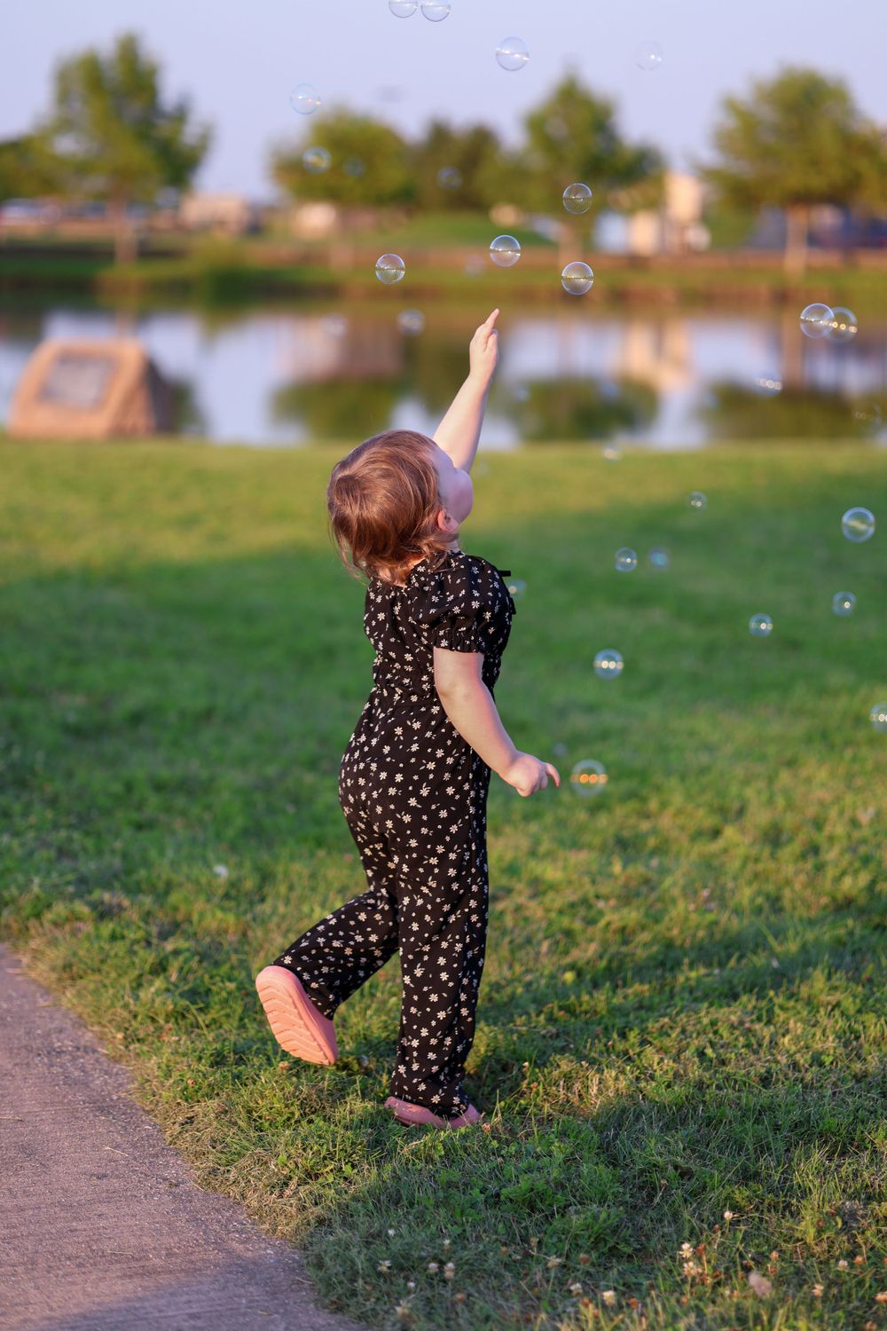 A small child in a polka dot dress reaches up to catch bubbles near a lake on a sunny day.
