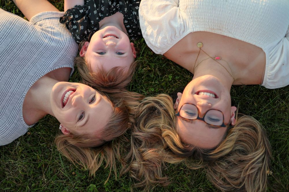Three people lie in the grass together smiling up at the camera during golden hour.