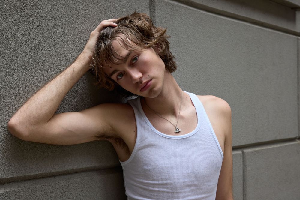 A person wearing a white tank top leans against a gray wall with hand resting on their head.