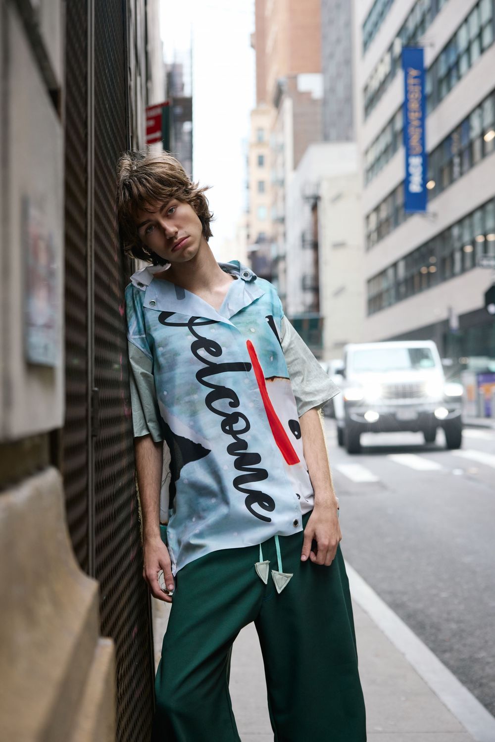 A person in a light blue graphic t-shirt and dark green pants poses casually against an urban building wall.