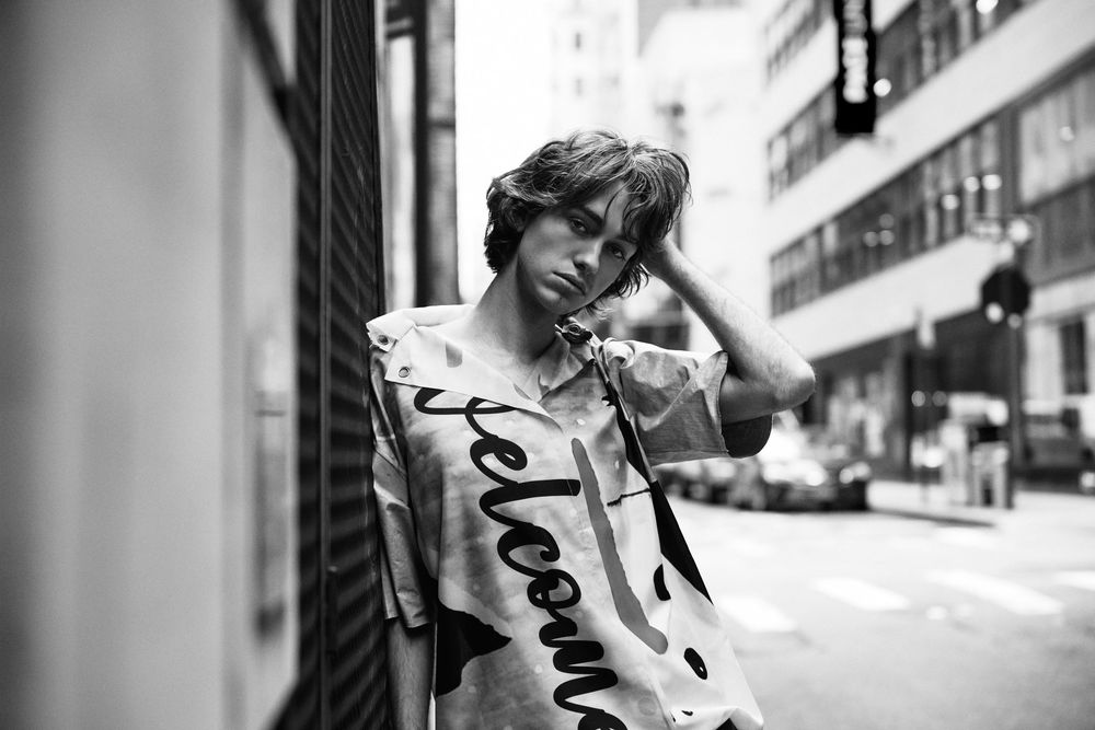 Black and white portrait of person leaning against wall wearing script text t-shirt on city street.