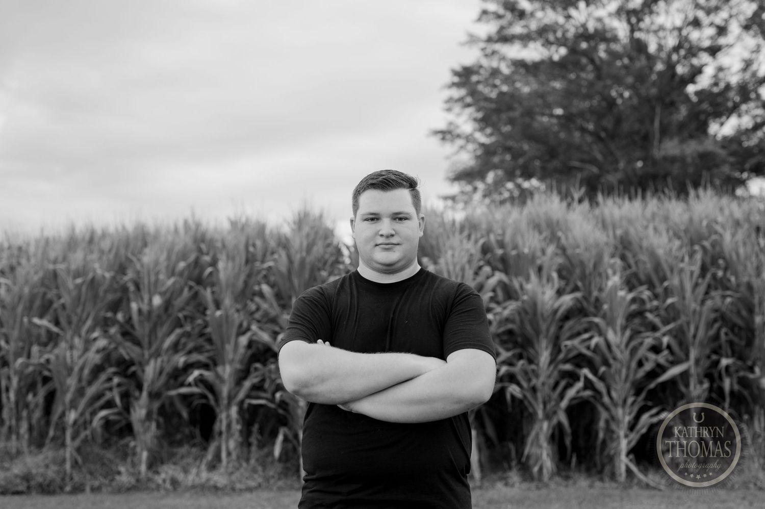 A person in a black shirt stands with crossed arms in front of a cornfield on an overcast day.
