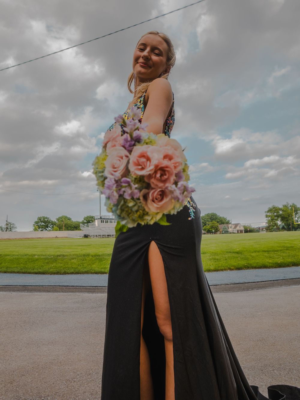 Formal black gown with floral beaded bodice and pink bouquet against cloudy sky.