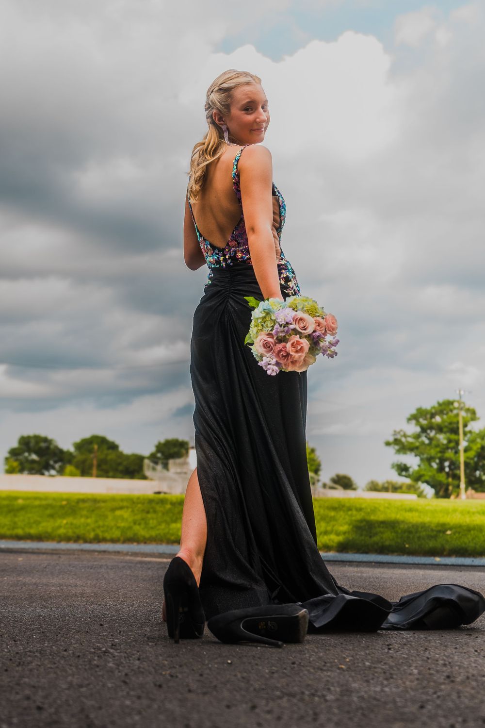 A woman in a black prom dress with floral details poses with a bouquet against a cloudy sky.