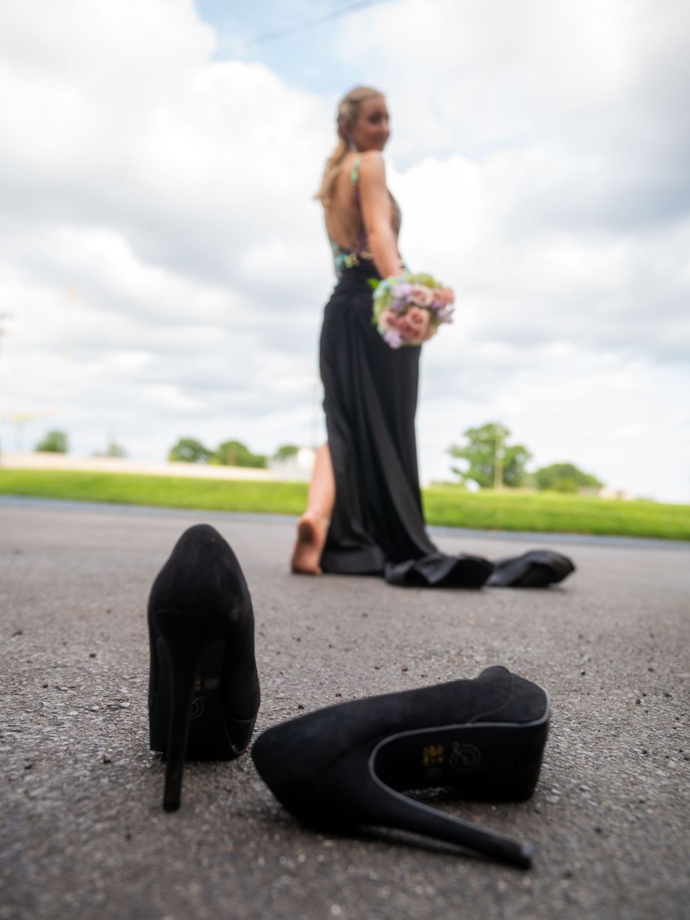 Black high heels on pavement with a formal dress in the background.