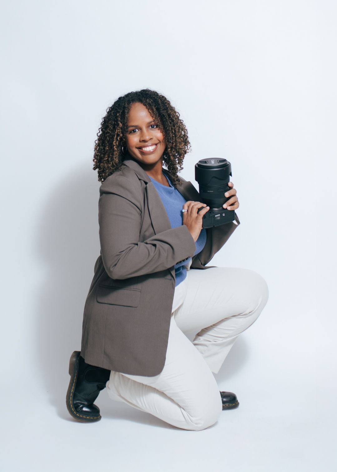 Professional photographer in a brown blazer kneeling with camera equipment against white backdrop.
