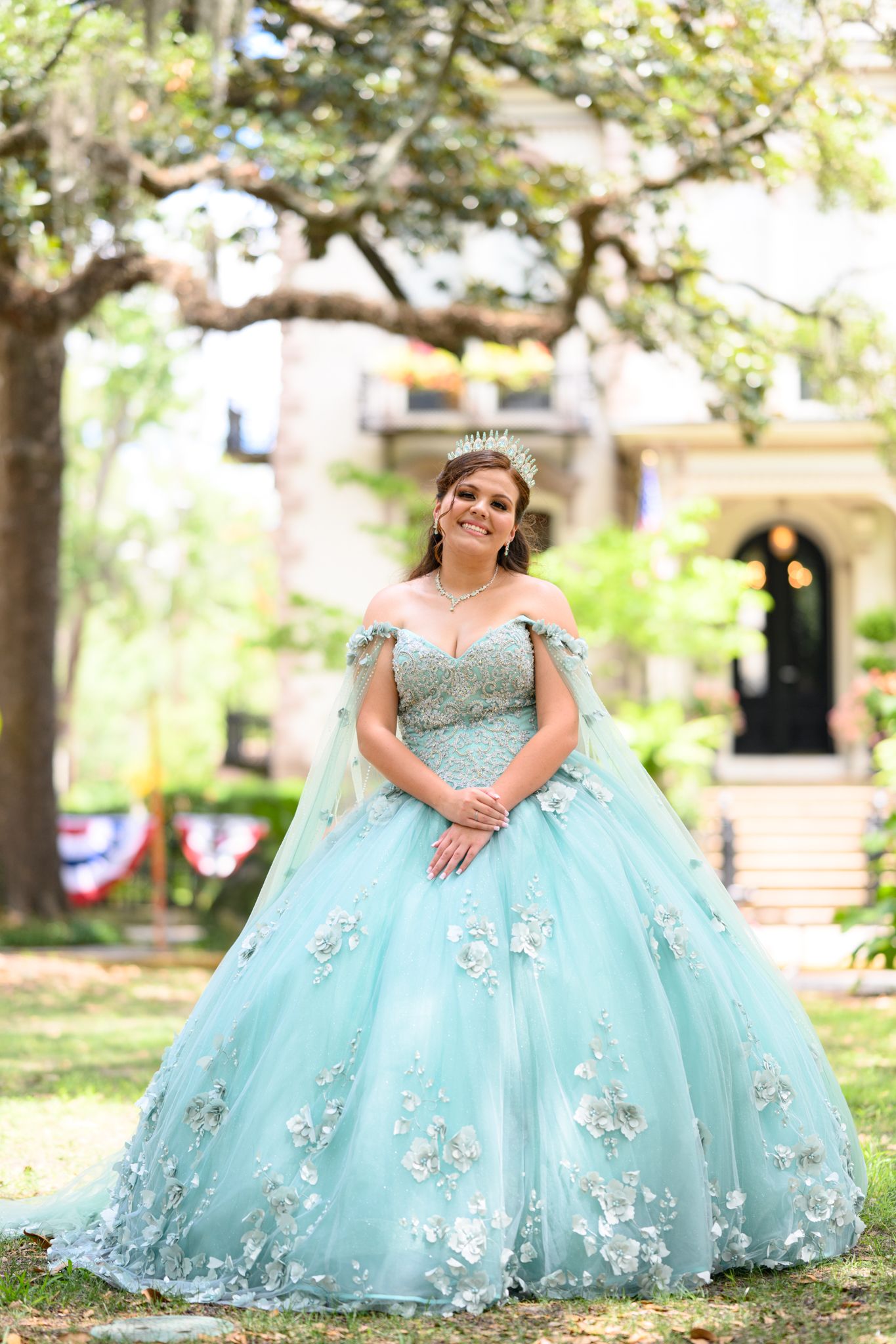 Person wearing light blue ball gown with floral appliques poses in garden setting.