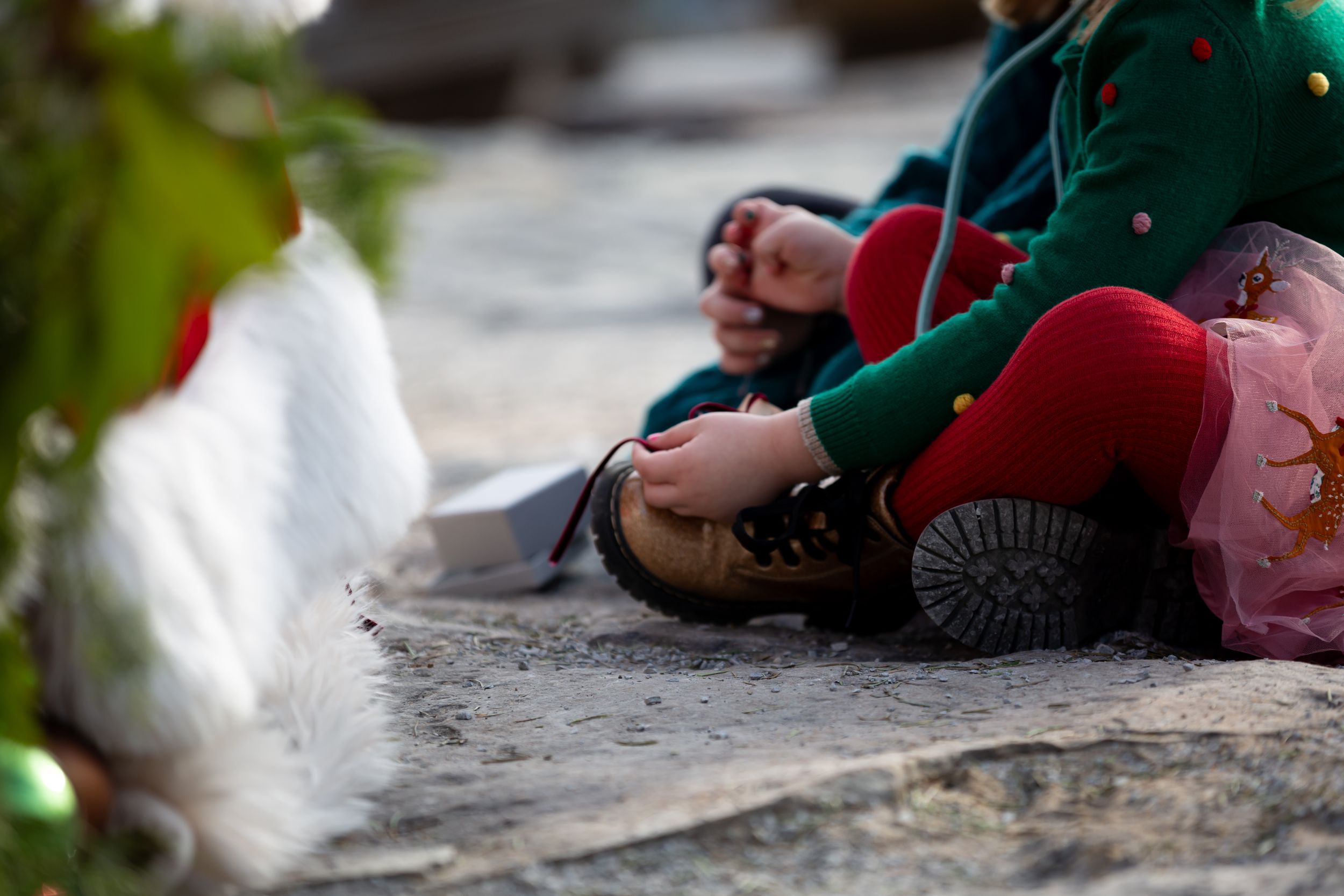 Child in green and red holiday attire sitting on ground near Christmas decorations.