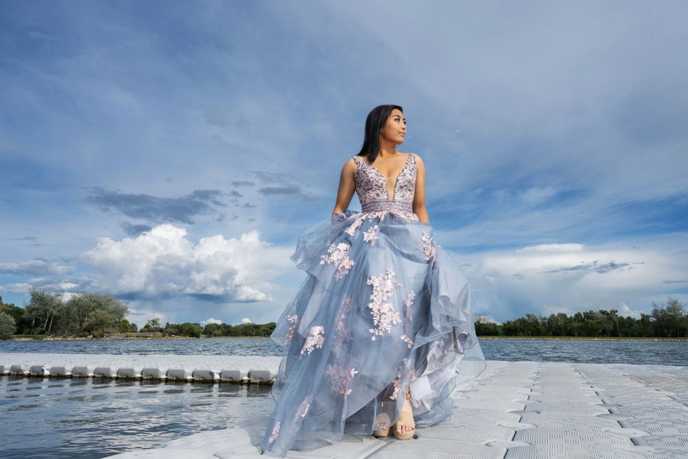Ethereal photoshoot of flowing blue dress on a dock over serene water under cloudy skies.