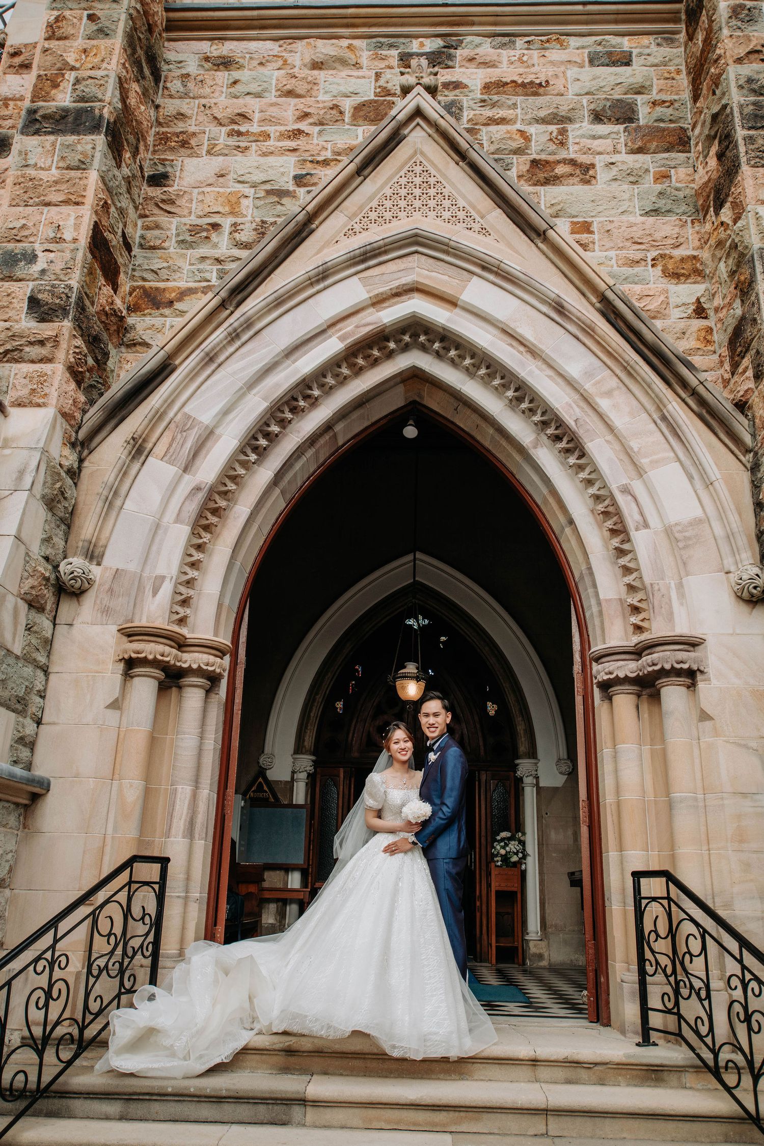 A grand church entrance frames a wedding portrait with ornate stone archways and iron railings.