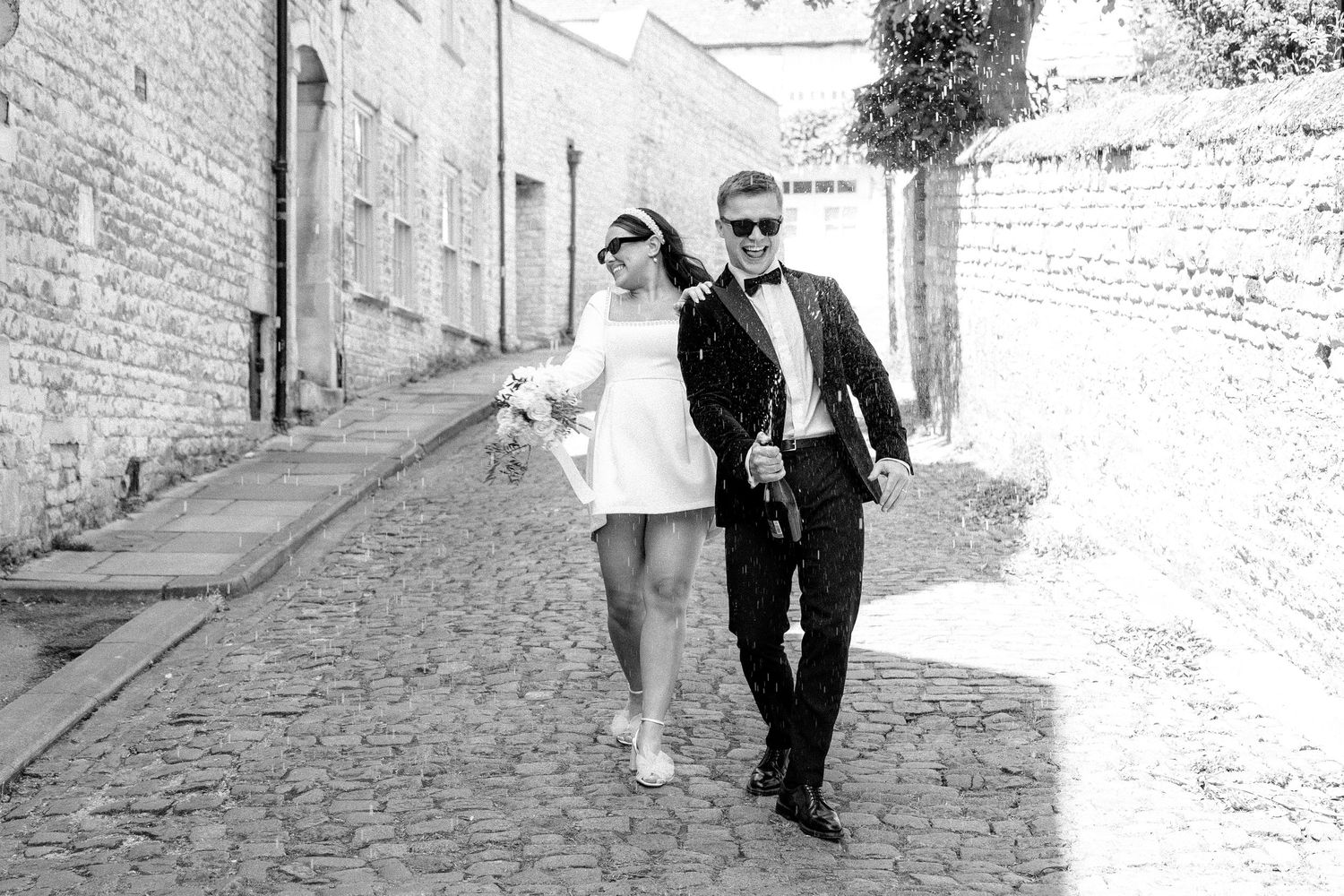 Black and white photo of a couple walking down a cobblestone alley in wedding attire.