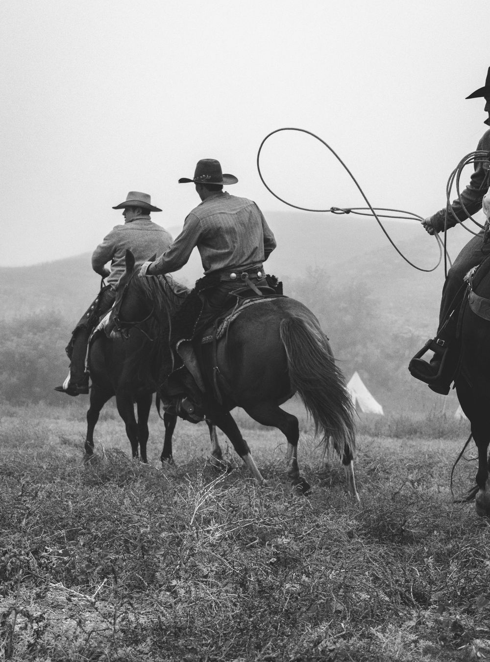 Riders on horseback galloping through misty grassland in black and white.