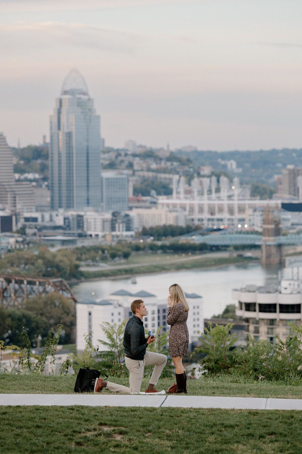 Two people sit on a grassy hill overlooking the Cincinnati skyline and Ohio River on a cloudy day.