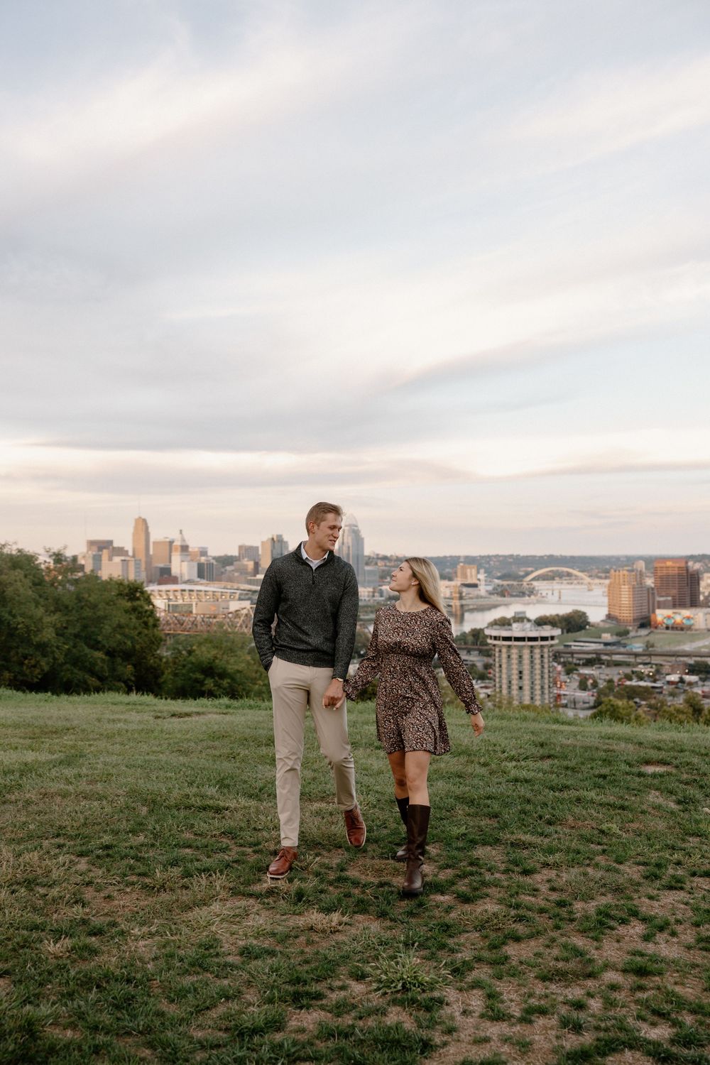 A couple stands on a hilltop overlooking a cityscape at sunset, with downtown buildings and bridges visible in the distance.