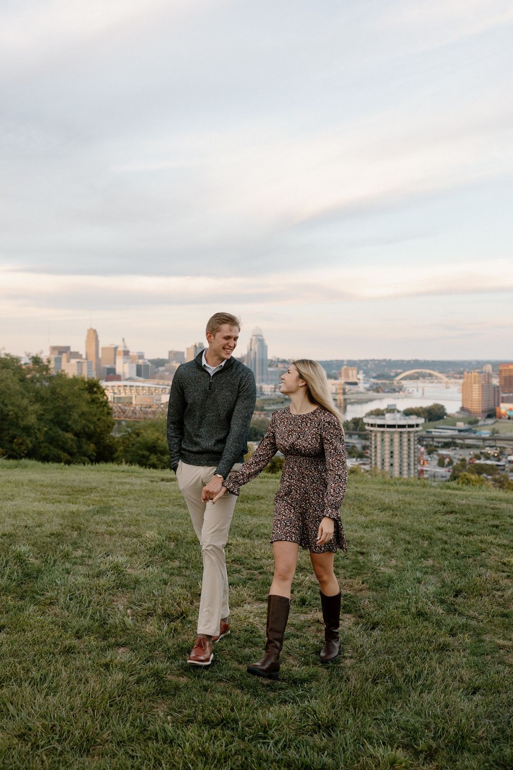 A couple shares a romantic moment on a grassy hilltop overlooking a city skyline during sunset.