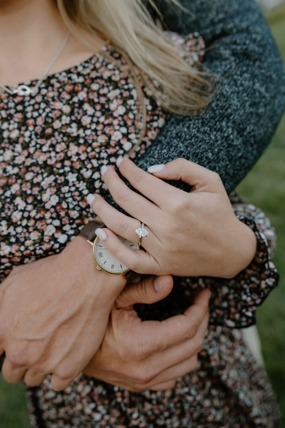 Close-up of hands showing an engagement ring against a floral dress and gray sweater.