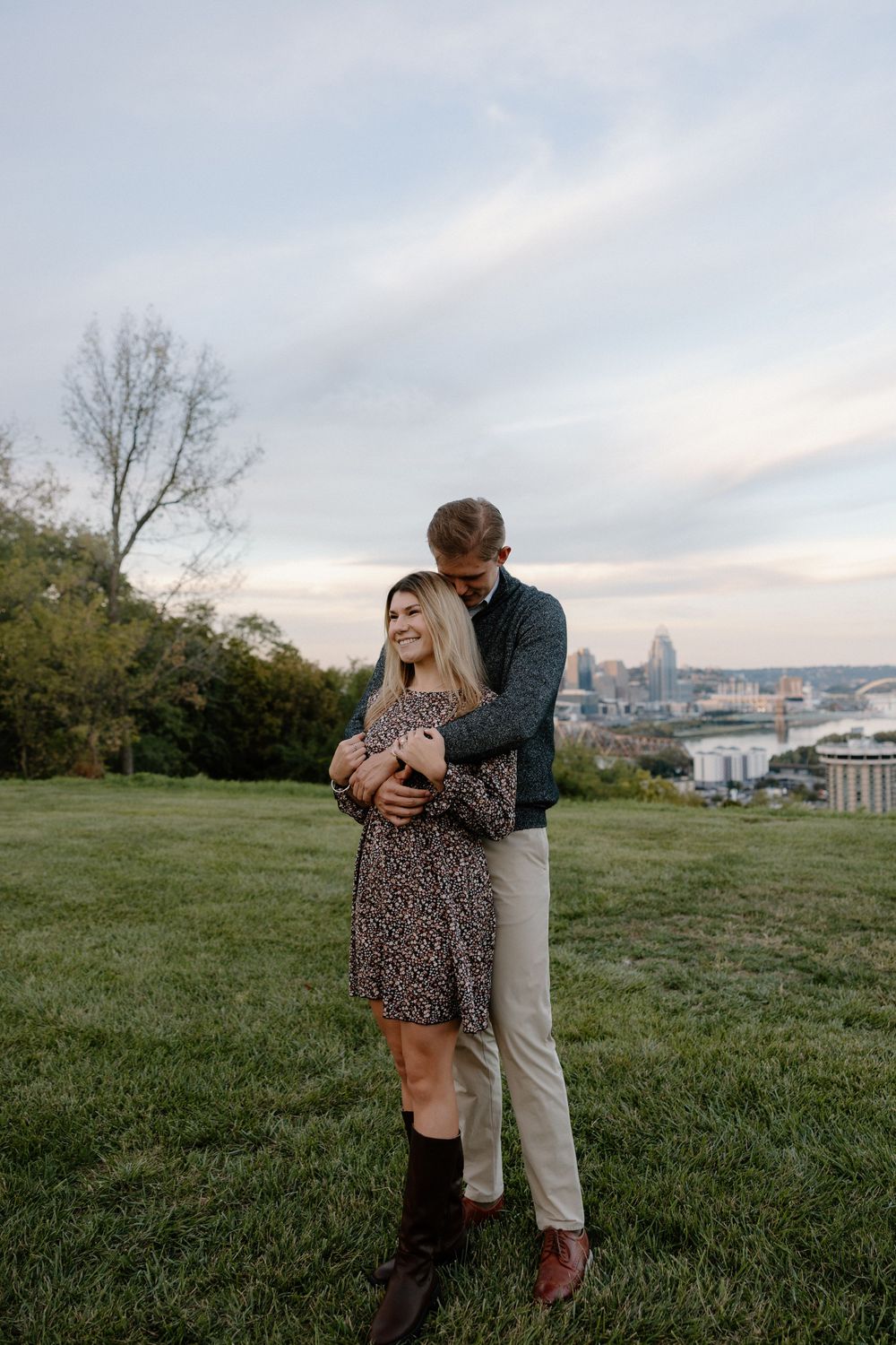 A couple shares a romantic moment on a grassy hilltop overlooking a city skyline during golden hour.