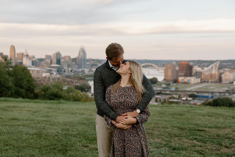 A couple embraces on a grassy hilltop overlooking a cityscape at sunset.