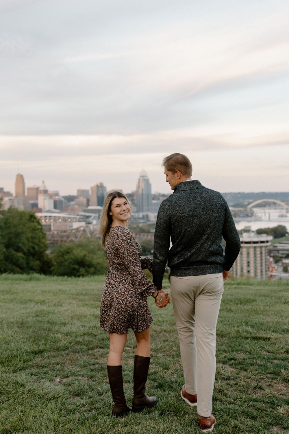 A couple walks hand in hand on a grassy hilltop overlooking a city skyline during sunset.