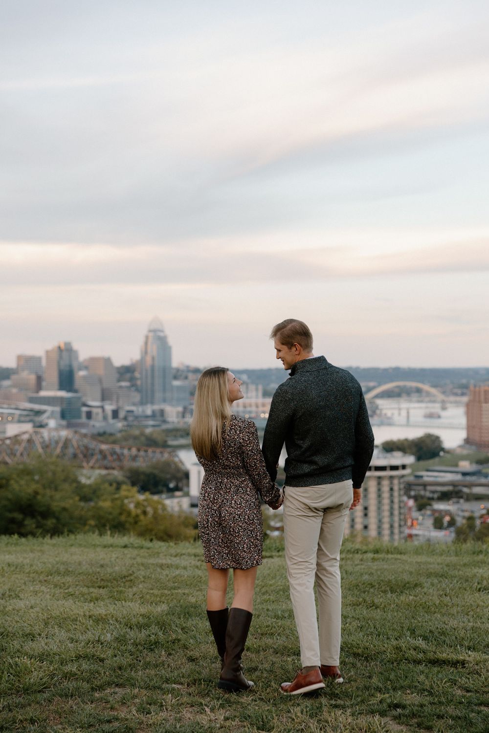 Couple walking on a hilltop overlooking a city skyline with bridges and buildings at sunset.